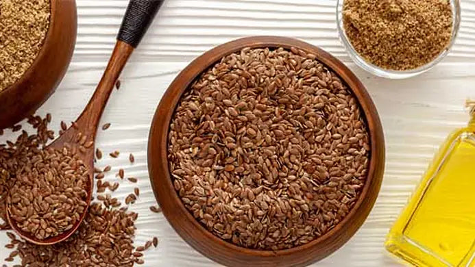 Wooden bowls of whole brown linseed and milled flaxseed, a spoon of seeds and a bottle of flaxseed oil on a white wooden table