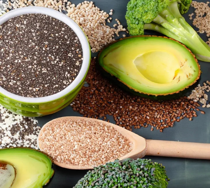 Chia seeds in a green bowl with halved avocados, broccoli, a wooden spoon of seeds and scattered seeds on a dark surface