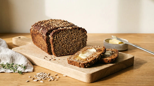 Sliced dark seeded protein loaf on an oak board, its dense crumb full of linseed and sunflower seeds, with two toasted buttered slices and a dish of butter